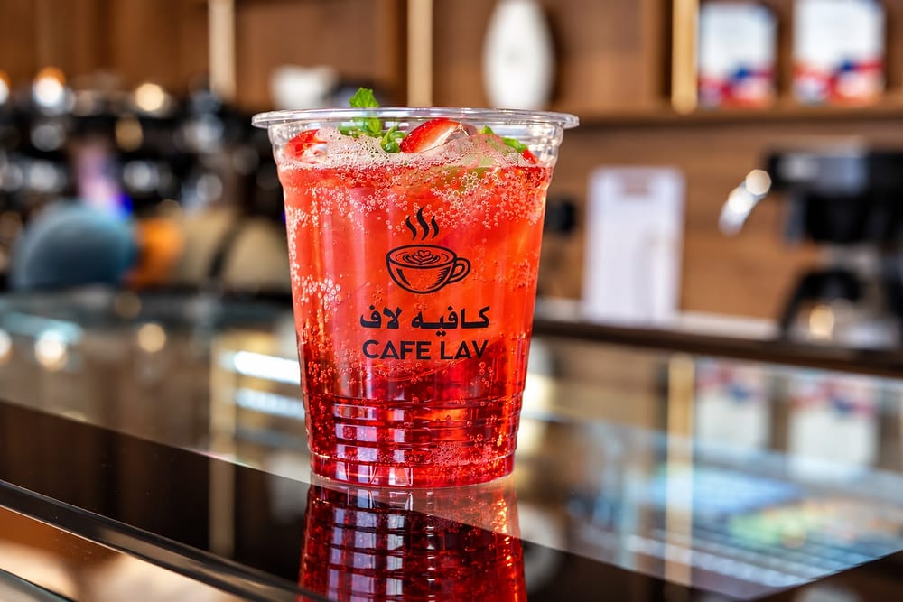 كوب مشروب أحمر مثلّج بالفراولة والنعناع يحمل شعار كافيه لاف ⁦CAFE LAV⁩ على منضدة زجاجية، وخلفه آلة القهوة والرفوف
